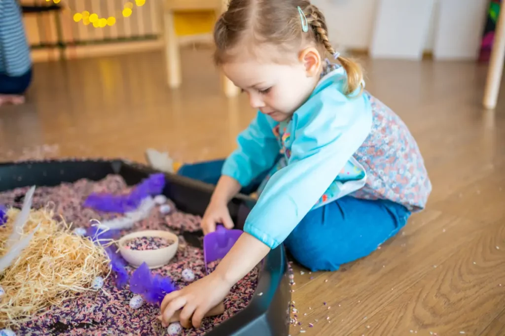 Dziewczynka szuka przedmiotów w baseniku, zabawy sensoryczne dla dzieci